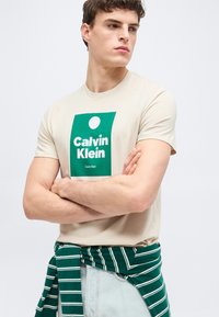 Νεαρός άνδρας με σταυρωμένα χέρια, φορώντας μπεζ μπλουζάκι Calvin Klein και πράσινο πουλόβερ με ρίγες δεμένο στη μέση, κοιτάζει πλάγια.