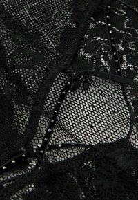 Bodysuit negro de encaje con patrones de red y florales, con acentos de cinta de satén y pequeños studs metálicos a lo largo de los bordes.