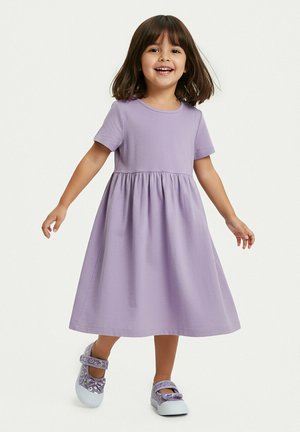 Lächendes junges Mädchen mit schulterlangen braunen Haaren, das ein lavendelfarbenes, kurzärmeliges Kleid und passende Schuhe trägt, steht mit leicht ausgestreckten Armen.