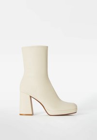 Botín en crema hasta el tobillo con una textura suave similar al cuero, punta cuadrada y un tacón alto y grueso. Presenta detalles mínimos de costura y un diseño elegante.