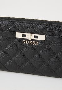 Schwarze, gestepelte Geldbörse aus Lackmaterial mit goldfarbenem Verschluss und Logo, das ein geprägtes Muster aufweist.