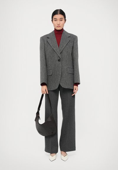 Blazer en laine sur mesure gris foncé sur un col roulé rouge, associé à un pantalon large gris et des escarpins blancs à bout pointu. Sac à main tissé noir.