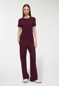 Bordowy t-shirt z krótkim rękawem w połączeniu z dopasowanymi szerokimi spodniami. Obie części wykonane z gładkiego, miękkiego materiału. Model stoi z rękami w kieszeniach.