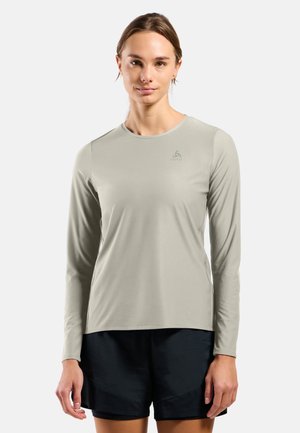 Femme portant un t-shirt de sport à manches longues gris clair et un short noir, debout devant un fond clair uni.