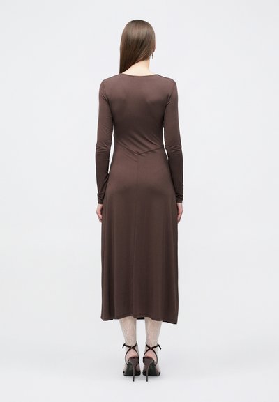 Άτομο που στέκεται με την πλάτη, φορώντας ένα καφέ μακρυμάνικο μίντι φόρεμα, λευκές δαντελωτές κάλτσες και μαύρα ψηλοτάκουνα σανδάλια, σε απλό φόντο.