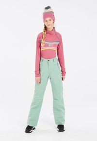 Aansluitend roze lange mouwen top met grijze en gele strepen, gecombineerd met losse mintgroene sneeuwbroek. Zwarte winterlaarzen en een roze gebreide muts met pompon.