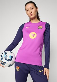FC Barcelona jersey in bright pink with blue long sleeves. Features Spotify logo, club crest, and is made of athletic fabric. Player holds a soccer ball.
