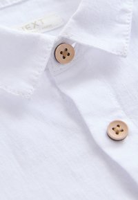 Chemise en lin blanc avec col, dotée de deux boutons en bois à quatre trous et d'un tissu légèrement texturé.