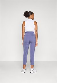 Polo sans manches blanche pour femme et pantalon de sport violet avec une coupe ajustée, longueur jusqu'aux chevilles et poches arrière. Les chaussures sont blanches avec des accents noirs.