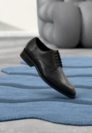 Zapato de vestir de cuero negro flotando sobre una alfombra azul ondulada con una silla acolchada gris al fondo.