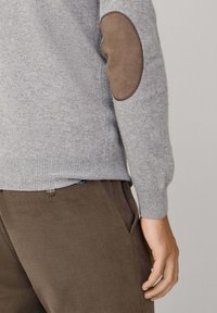 Primer plano de una persona con un suéter gris con un parche marrón en el codo y pantalones marrones, con la mano descansando al lado del muslo.