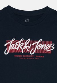 Námornícky modré tričko s bielym a červeným logom "Jack & Jones" a textovým detailom, umiestnené na pruhovanom pozadí.