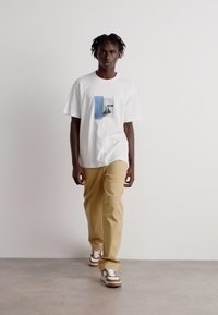 Hvit t-skjorte med grafisk trykk, beige bukser og sneakers med brune detaljer. Modellen står mot en ensfarget bakgrunn som viser antrekket.