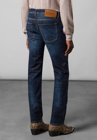 Dark blue denim jeans with a straight cut, featuring two back pockets and a tan leather branding patch on the waistband. Textured surface.