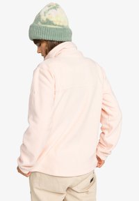 Mikina z fleece materiálu v jemnej rúžovej farbe, s vysokým golierom a dlhými rukávmi. Má šev na chrbte a diskrétnu značku na ľavom rukáve.