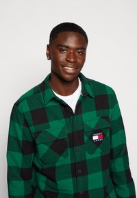 Camicia di flanella a quadri verdi e neri con due tasche sul petto, dotata di una patch con logo. Design con bottoni, maniche lunghe e colletto.