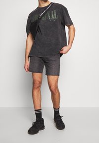 Black acid-wash oversized t-shirt with green "CRIMINAL" text, paired with dark denim shorts. Black sneakers and striped socks. Black crossbody bag.