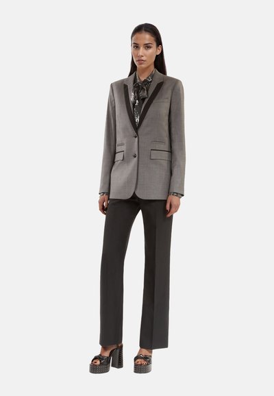 Maßgeschneiderter grauer Blazer mit schwarzen Revers, kombiniert mit einer bedruckten Bluse und schwarzen Schlaghosen. Verfügt über klobige schwarze Plateau-Schuhe.