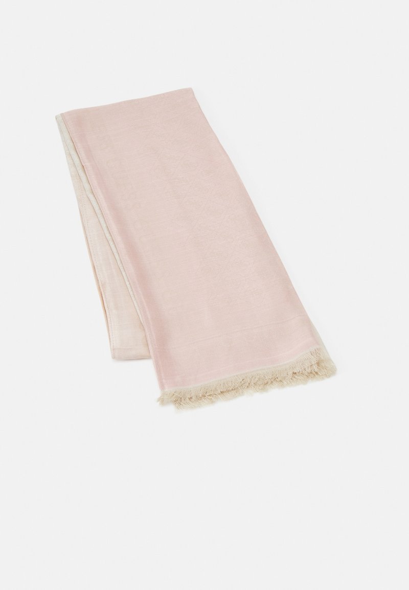 Serviette rose pâle douce pliée avec des bords à franges beige posée sur un fond blanc uni.