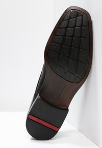 Semelle de chaussure noire avec une surface en caoutchouc texturée, des motifs rectangulaires antidérapants et un accent marron au talon avec le nom de la marque embossé.