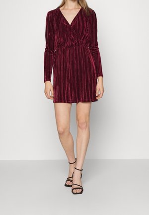 Vrouw draagt een kort bordeauxrode fluwelen jurk met lange mouwen en een V-hals, gecombineerd met zwarte sandalen met hakken en bandjes.