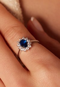 Saffier ring met een centrale blauwe steen omringd door kleine heldere edelstenen, gezet op een verzilverde band met een gepolijste afwerking.