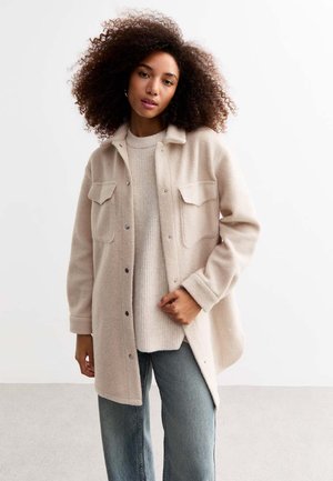 Beige wool blend shirt jacket with two chest pockets, button front, and relaxed fit, layered over a light textured sweater and denim jeans.