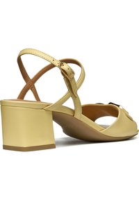 Sandalias de tacón cuadrado de cuero amarillo con diseño de tiras cruzadas, punta abierta y detalle decorativo de moño en la parte frontal. Acentos marrones visibles.