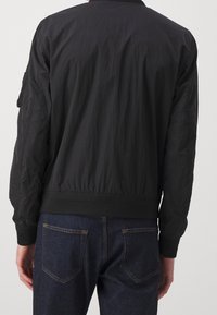 Giubbotto bomber nero con finitura testurizzata, polsini e orlo a costine, e dettagli a toppa sulle maniche; abbinato a jeans di denim scuro.