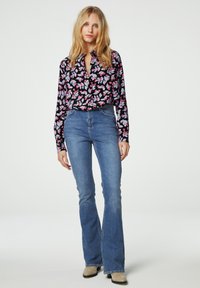 Zwarte blouse met paars en roze bloemenpatroon, gecombineerd met lichtblauwe flared jeans. De gladde textuur en de getailleerde pasvorm zorgen voor een casual uitstraling.