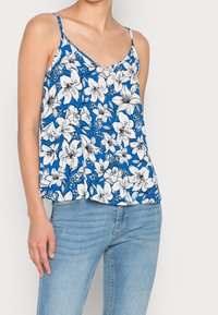 Débardeur floral bleu avec des motifs de lys blancs, design en V, et fines bretelles, associé à un jean en denim bleu clair.