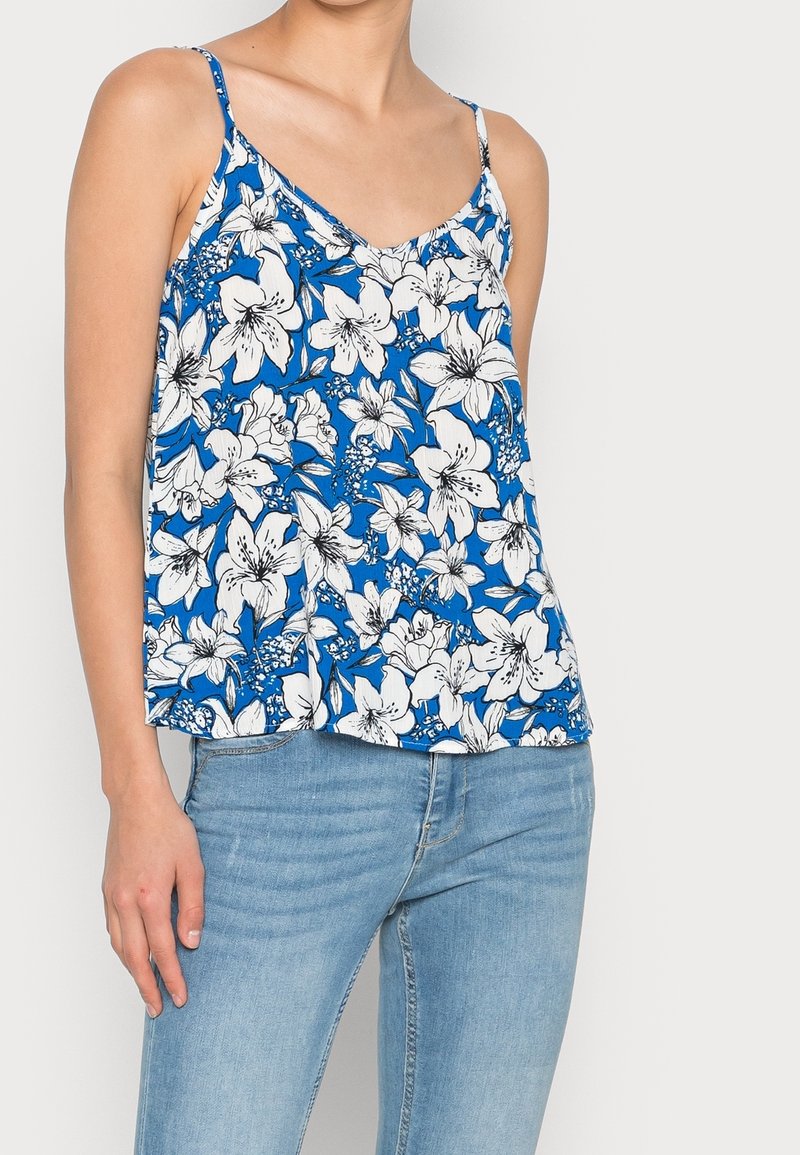 Débardeur floral bleu avec des motifs de lys blancs, design en V, et fines bretelles, associé à un jean en denim bleu clair.