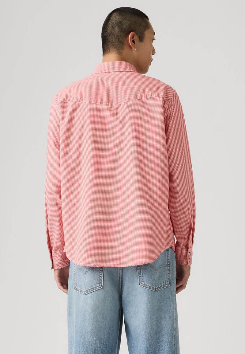 Uomo che indossa una camicia rossa a quadri a maniche lunghe e jeans azzurri chiari, di spalle su uno sfondo semplice grigio chiaro.