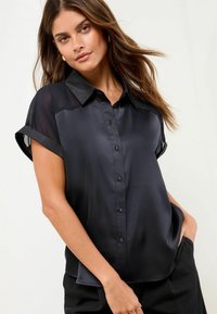 Schwarzes Satin-Hemd mit kurzen, umgeschlagenen Ärmeln, einem strukturierten Kragen und einer glatten Textur. Verfügt über eine lässige Passform und einen dezenten Glanz.