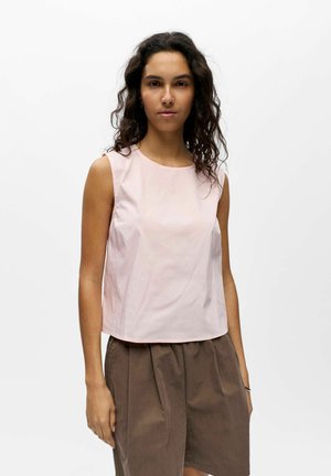 Femme aux longs cheveux bouclés, portant un haut rose clair sans manches et un pantalon marron ample, debout devant un fond blanc uni.