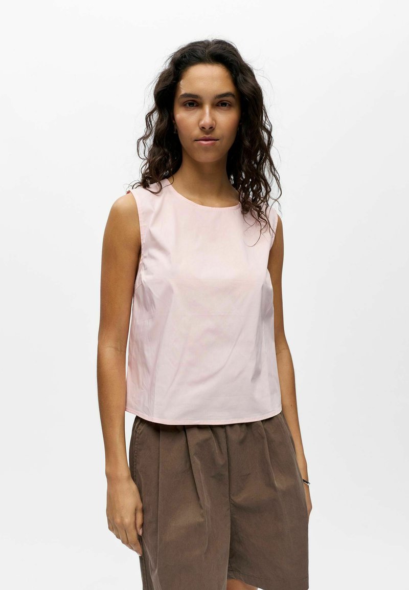 Femme aux longs cheveux bouclés, portant un haut rose clair sans manches et un pantalon marron ample, debout devant un fond blanc uni.