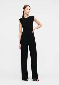 Γυναίκα με μαύρη ολόσωμη φόρμα χωρίς μανίκια με φαρδιές παντελόνες και κρεμαστά σκουλαρίκια, στάση με το ένα χέρι στη μέση.