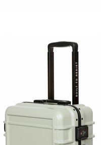 Tvrzený kufr světle šedé barvy s černou teleskopickou rukojetí, černými kovovými detaily a kombinovaným zámkem. Text čte: "NAVRHNUTO, ABY VYDRŽELO."