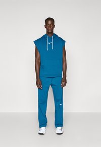 Haut sans manches bleu avec poche kangourou et cordons blancs, associé à un pantalon bleu assorti avec un accent de logo blanc.