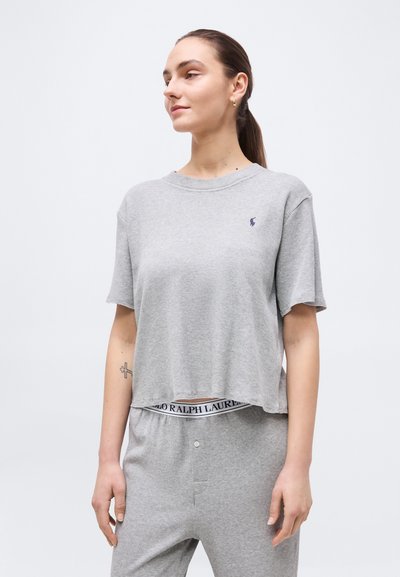 Γυναίκα που φοράει ανοιχτό γκρι κοντομάνικη μπλούζα και ταιριαστό παντελόνι, με ορατή λευκή ζώνη μέσης με την επωνυμία "Polo Ralph Lauren", στέκεται μπροστά σε απλό φόντο.
