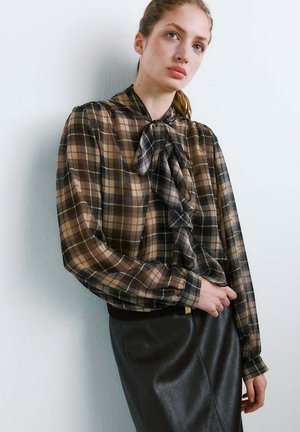 Kobieta w brązowej, kratkowanej bluzce z kokardą na kołnierzyku oraz czarnej skórzanej spódnicy, stojąca przy białej ścianie.