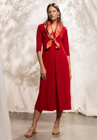 Robe rouge longueur genou avec un col en V et des manches trois-quarts, ornée d'une écharpe à motifs orange nouée au cou et de sandales dorées à brides.