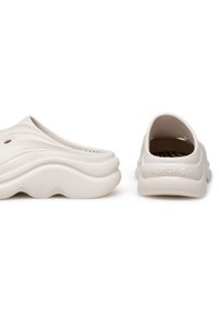 Cizme albe din material plastic cu un design ondulat, spate deschis și găuri de ventilație. Textură netedă cu o talpă groasă, având un branding subtil.