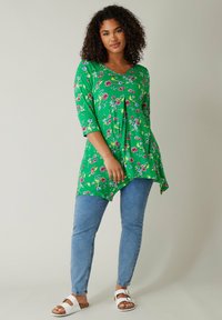 Grüne Blumentunika mit V-Ausschnitt, dreiviertel Ärmeln und asymmetrischem Saum, kombiniert mit hellblauen Skinny-Jeans und weißen Sandalen.
