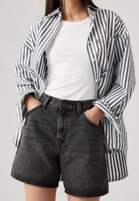 Camisa a rayas de botones en navy y blanco, combinada con pantalones cortos de mezclilla negra de cintura alta. Sencilla camiseta sin mangas blanca de canalé debajo de la camisa.