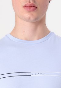 Gaiši zils kokvilnas T-krekls ar apaļu kaklu, uz kura ir melnbalta "TOMMY JEANS" logo un divas izliekta līnijas pāri krūtīm.