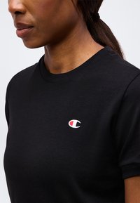 Γυναίκα φορώντας μαύρο T-shirt με στρογγυλή λαιμόκοψη και μικρό κόκκινο και λευκό λογότυπο Champion στο αριστερό στήθος, με τα μαλλιά της δεμένα σε αλογοουρά.