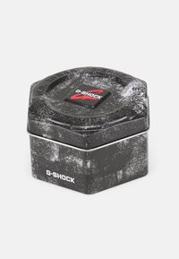 Conteneur en métal hexagonal G-SHOCK, noir avec une finition texturée, présentant un logo rouge sur le dessus et des éclats blancs sur la surface.
