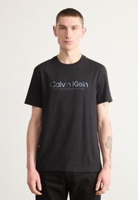 Sort bomulds T-shirt med rund halsudskæring og korte ærmer. Forsiden har "Calvin Klein" logoet i blå og metal nuancer.