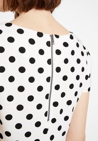 Bílé šaty s černými puntíky, krátké rukávy a viditelný zadní zip. Tkanina vypadá hladce s mírně texturovaným vzorem.